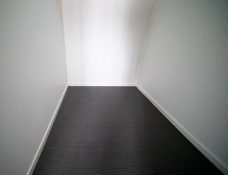 Empty narrow hallway with white walls and dark carpet, leading to a bright doorway.