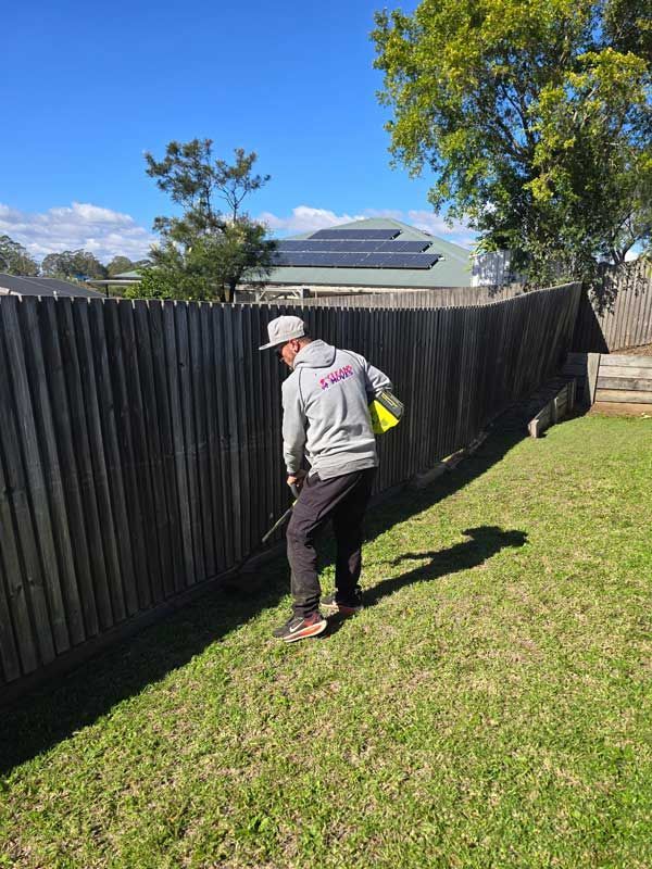 Person walking along a wooden fence in a sunny grassy yard with a whipper snipper.