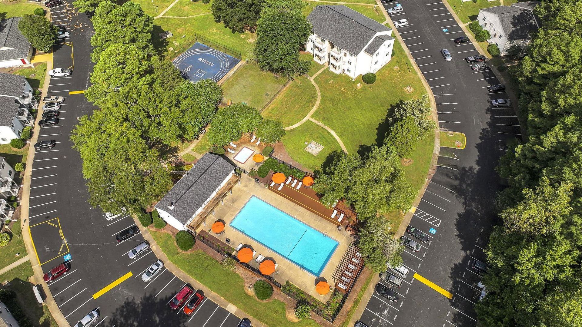 Aerial view of an apartment complex with a pool, playground, and parking. Green trees and blue pool.