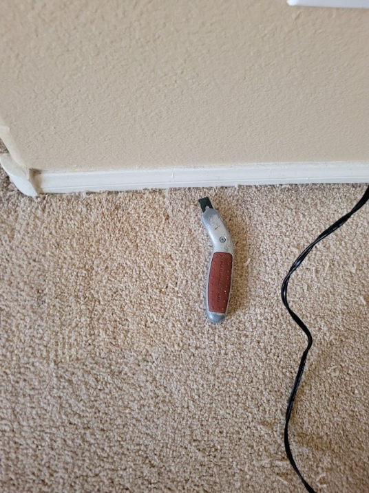Utility knife on beige carpet beside white baseboard and a black cord.