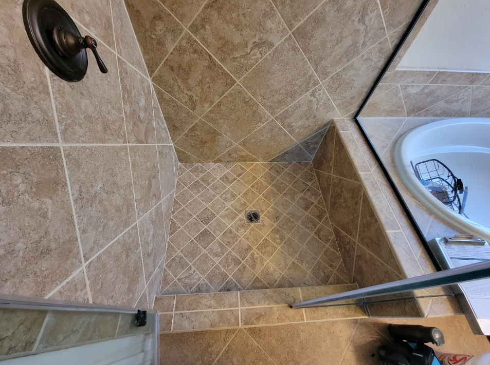 Shower stall with beige tile floor, glass door, and visible drain next to a sink.
