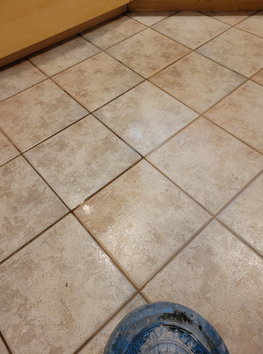 Tiled kitchen floor with beige tiles and grout lines, seen from above, with a blue bucket at the bottom edge