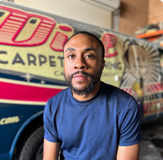 Man in a blue shirt seated in front of a colorful carpet cleaning truck sign
