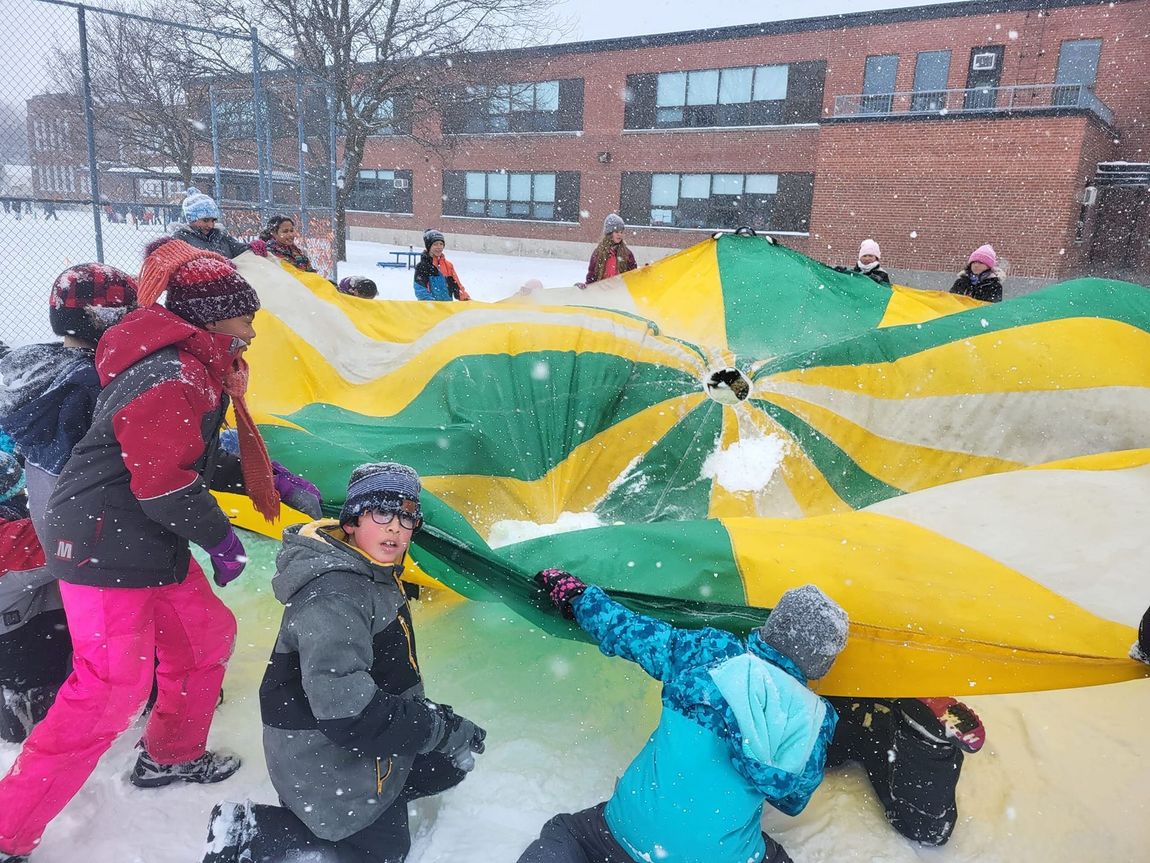 Children in winter coats hold a giant yellow-and-green parachute in a snowy schoolyard