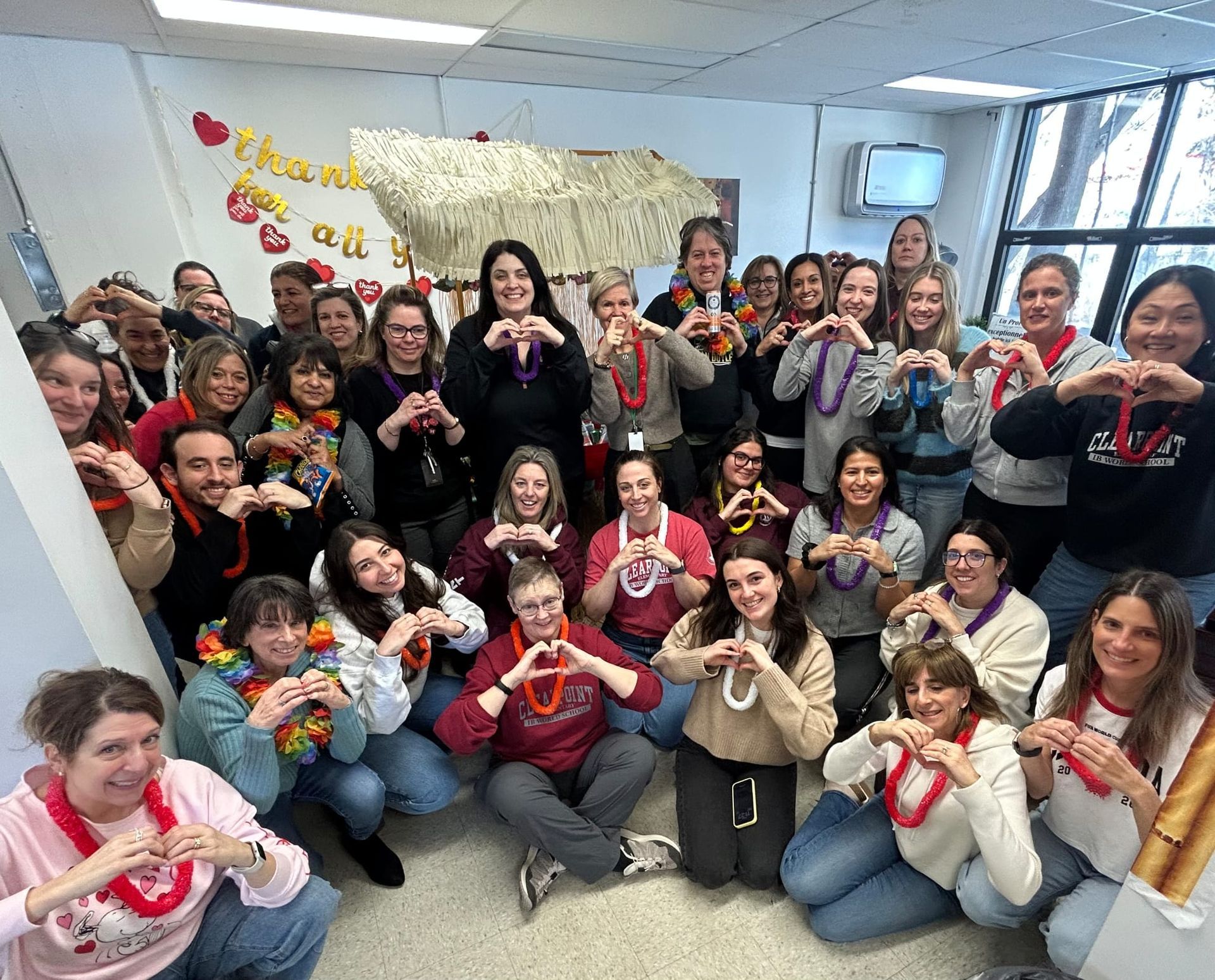 Large group posing indoors, smiling and holding red heart cutouts, with a festive Valentine’s Day wall display