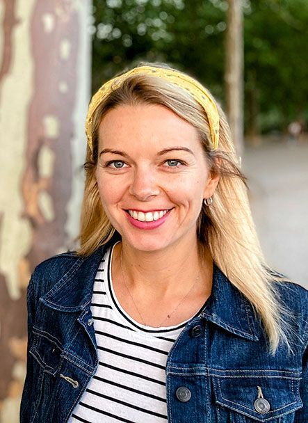 Smiling blonde woman in a denim jacket and striped shirt outdoors with blurred greenery behind her