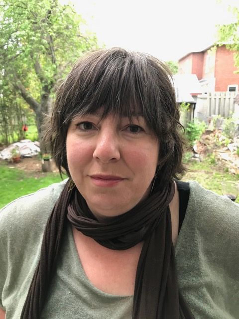 Woman outdoors in a garden, wearing a green top and dark scarf, with trees and a house behind her.