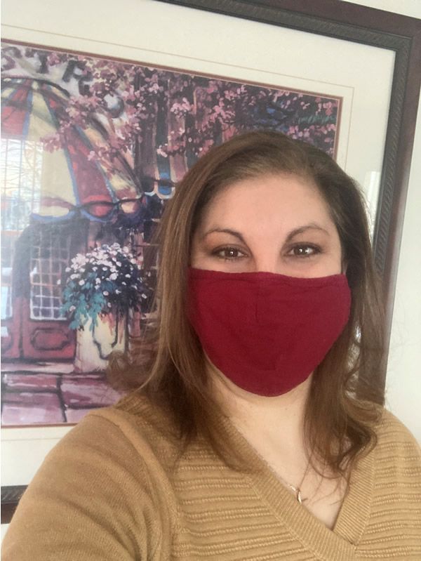Person wearing a red face mask and tan sweater stands in front of a framed floral painting.