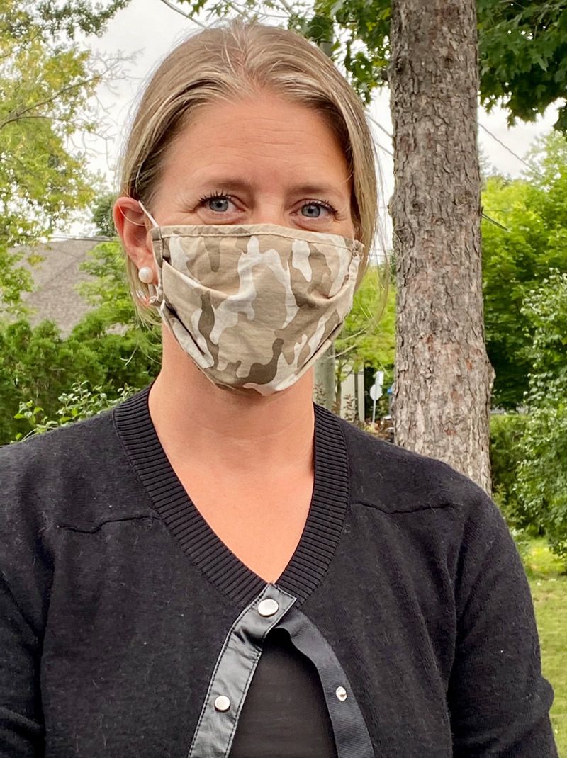 Woman wearing a camouflage face mask and black sweater outdoors beside a tree.