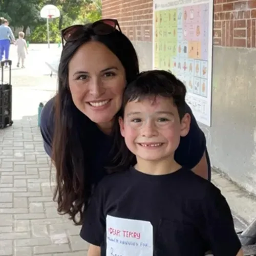 Smiling woman leaning beside a smiling child in a school hallway, with posters on the wall.