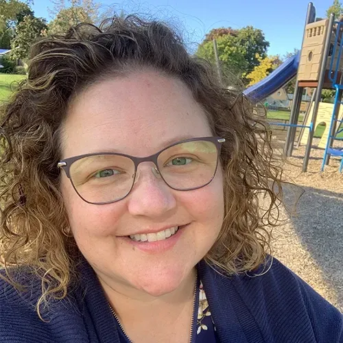 Smiling person with curly hair and glasses at a playground on a sunny day