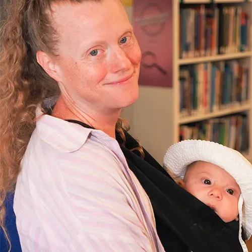 Person holding a baby in a black sling, both wearing white hats, in a room with bookshelves.