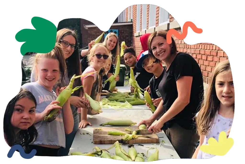 Group gathered around a table husking corn outdoors, smiling and holding green corn husks.