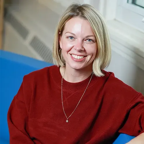 Smiling blonde woman in a red sweater standing indoors against a blue and white background