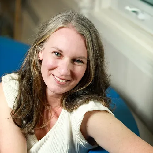 Smiling person taking a selfie indoors, wearing a cream top and sitting by a blue chair.