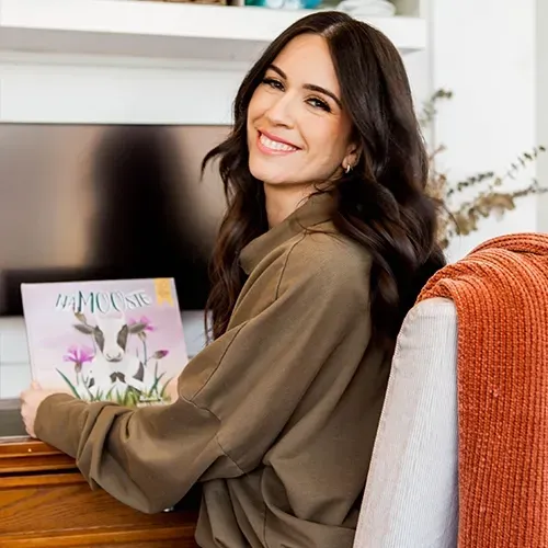 Smiling woman holding a floral card at a desk, with a TV and orange throw blanket in a cozy room.