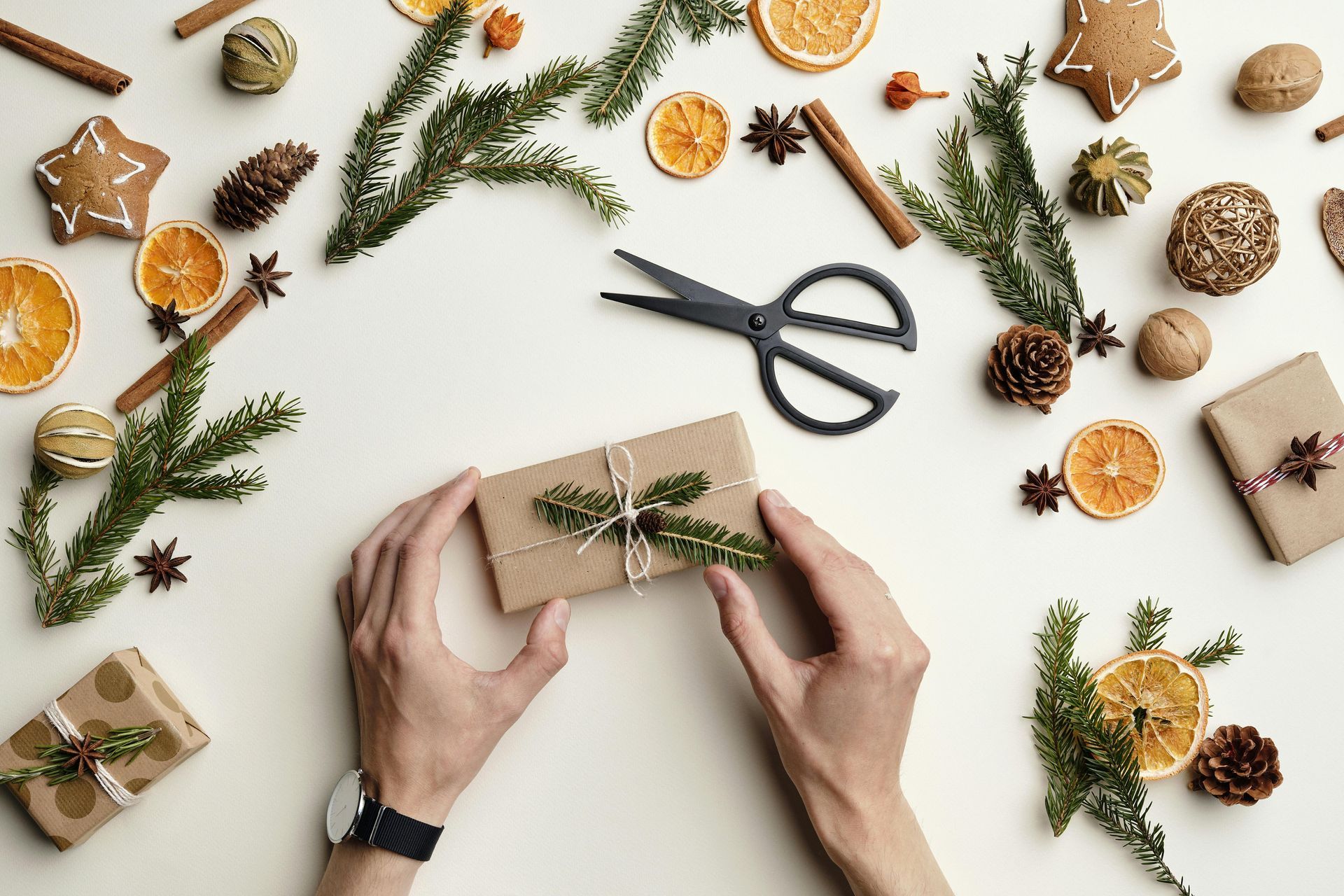 Hands wrapping a kraft gift box with twine, surrounded by pine sprigs, scissors, and dried oranges on a white table