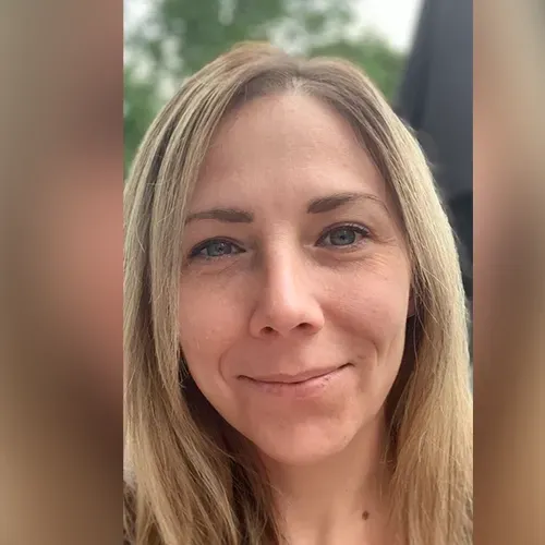 Close-up selfie of a smiling blonde woman outdoors with a blurred green background