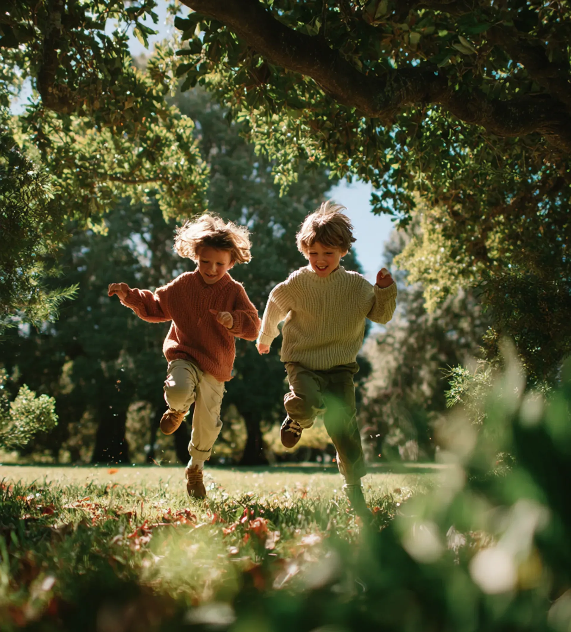 Two children running through a sunlit park beneath trees, one in orange and one in green.