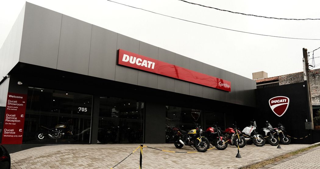 Uma motocicleta vermelha estacionada na calçada, ao lado de dois homens conversando, perto de prédios modernos.