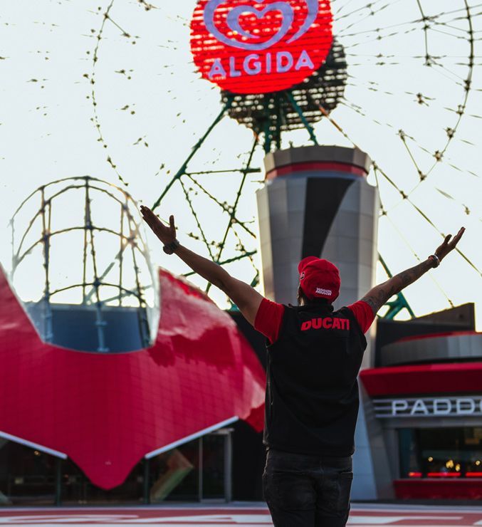 Pessoa vestindo uma camisa da Ducati com os braços erguidos em frente a uma roda-gigante com uma placa da Algida.