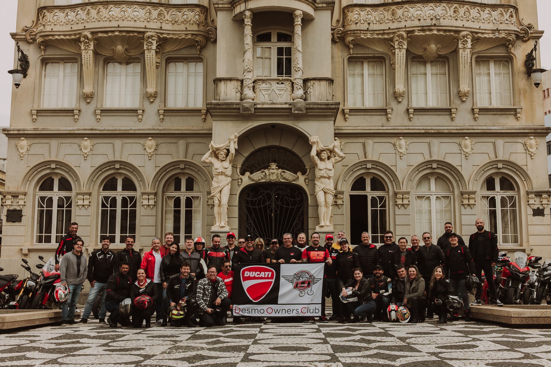 Um grande grupo posa e comemora em frente a um logotipo vermelho da Ducati na parede.