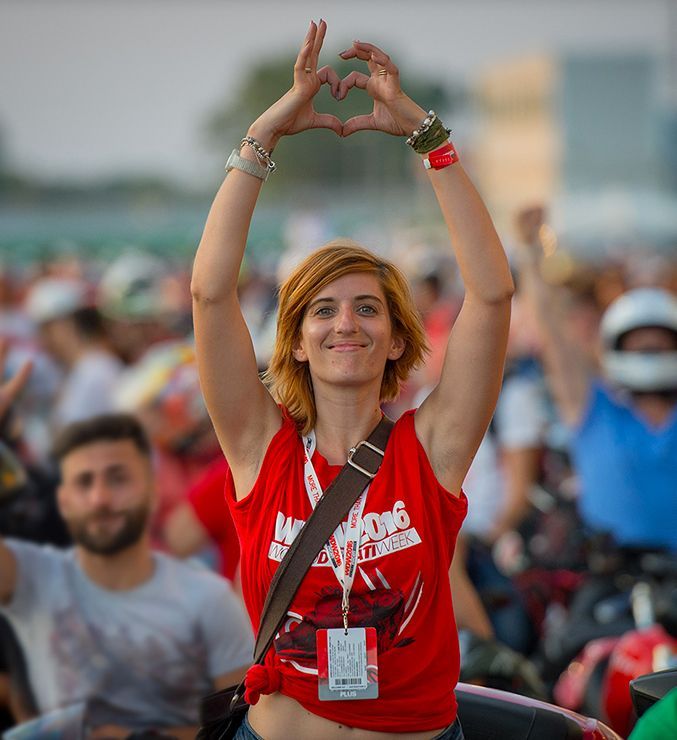 Mulher de camisa vermelha fazendo um coração com as mãos em um evento ao ar livre com grande público.