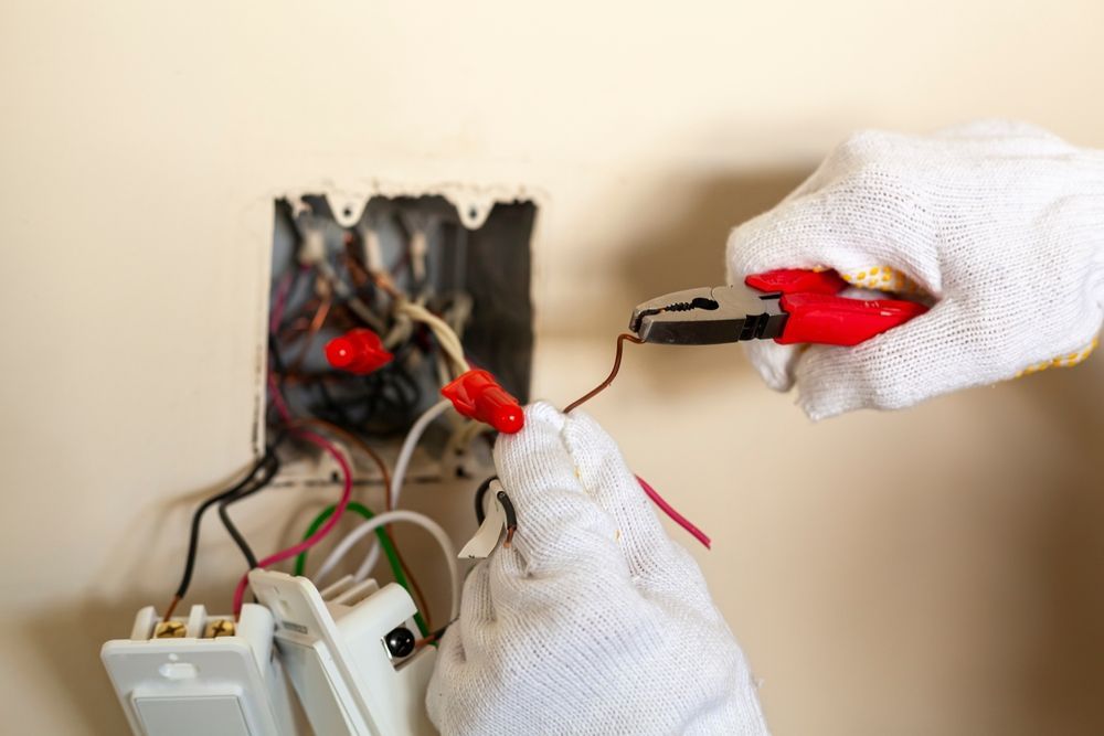 A pair of gloved hands uses pliers to work on electrical wiring in a wall box.