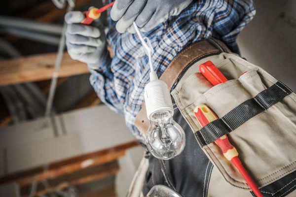 An electrician in a plaid shirt and tool belt holds a light bulb assembly while working in a construction setting.