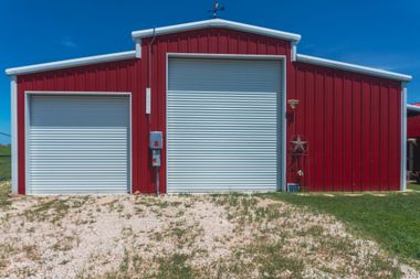 Red metal barn with white roll-up doors on a gravel lot under a blue sky