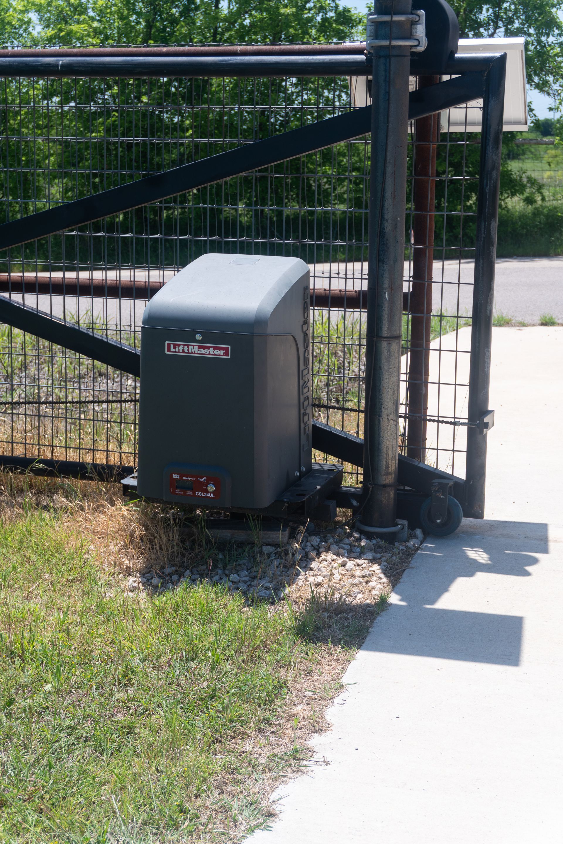 Gray trash bin beside a black metal gate on a sidewalk near a grassy area