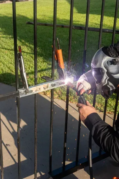 Person welding a black metal gate outdoors, with bright sparks and a grassy yard behind it