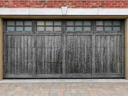 Weathered gray wooden garage doors beneath a red brick wall and concrete driveway
