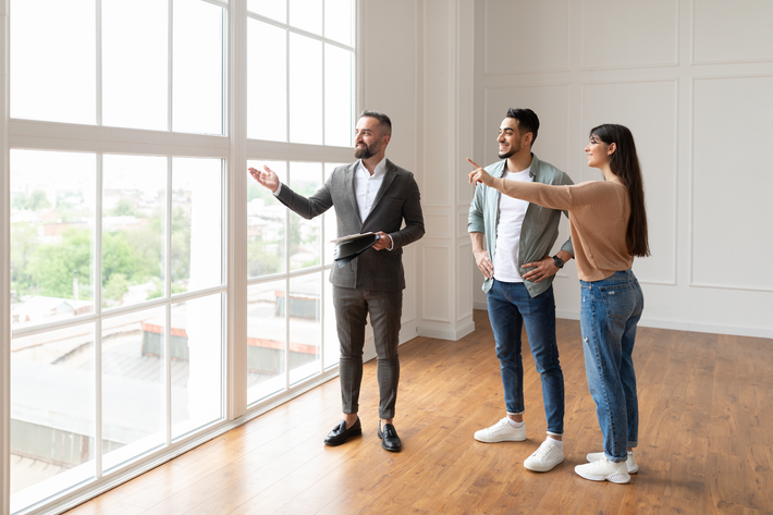 Three people touring a bright empty room, one gesturing toward large windows.