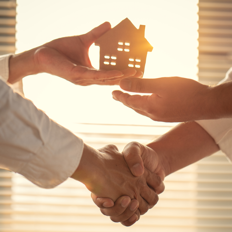 Hands passing a small house model over a handshake in warm sunlight