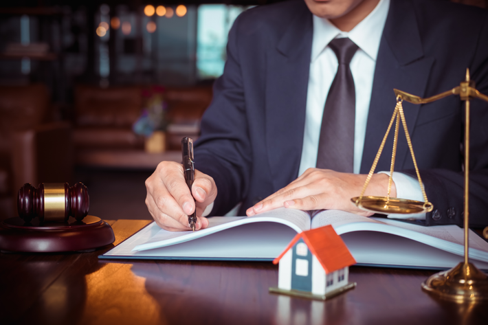 Lawyer writing in a ledger beside a gavel, scales of justice, and a small house model on a desk