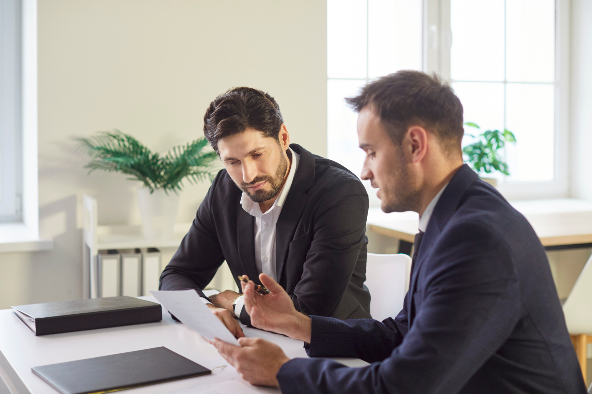Two businessmen reviewing documents at a bright office desk with a tablet and folders.