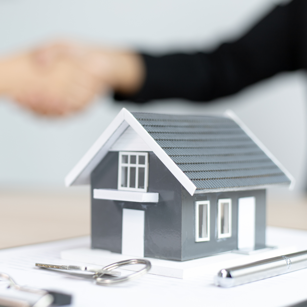 Model house beside keys and pen, with blurred handshake in the background