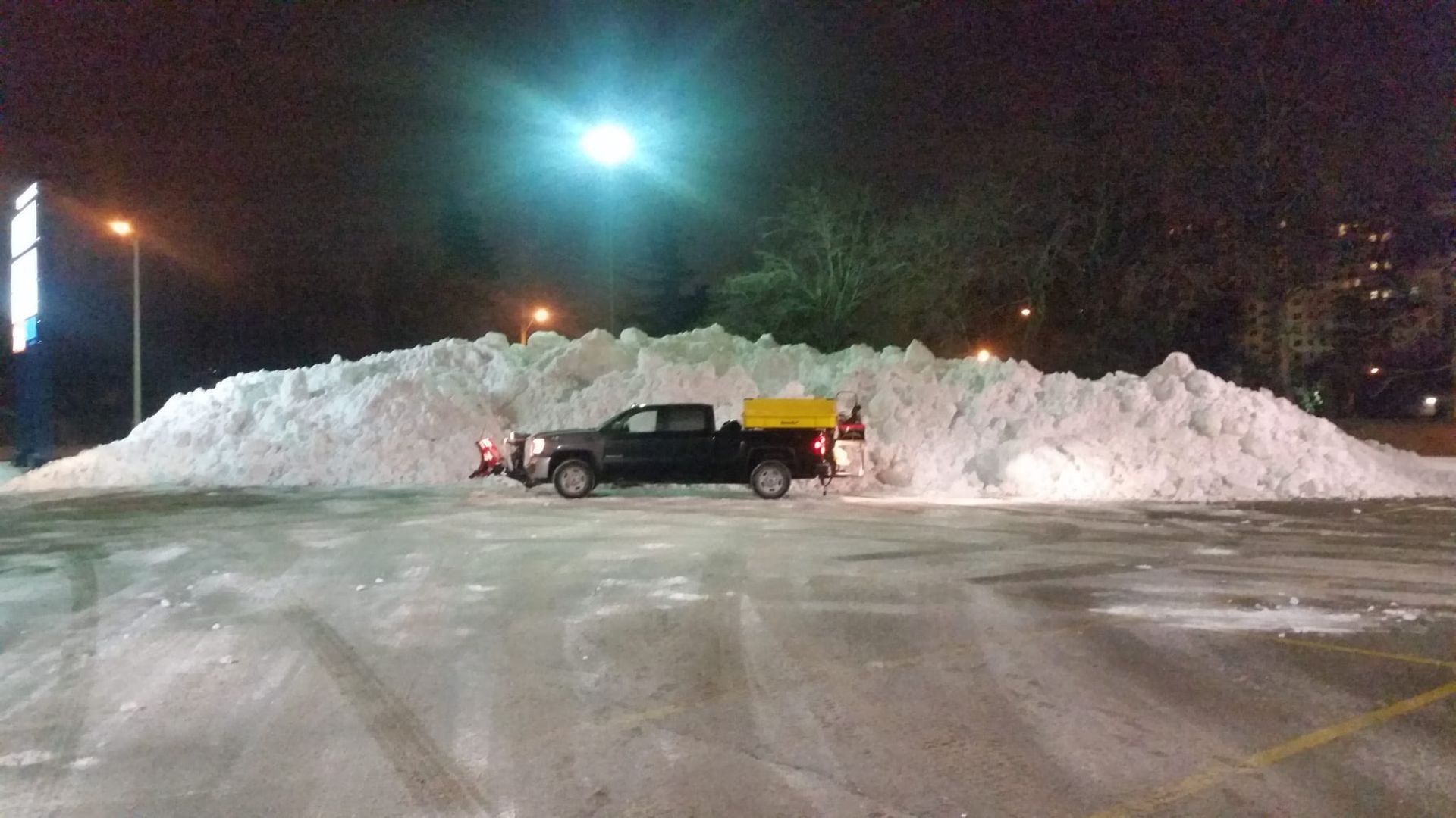 A black truck parked in front of a huge snow pile in a parking lot at night, under floodlights.
