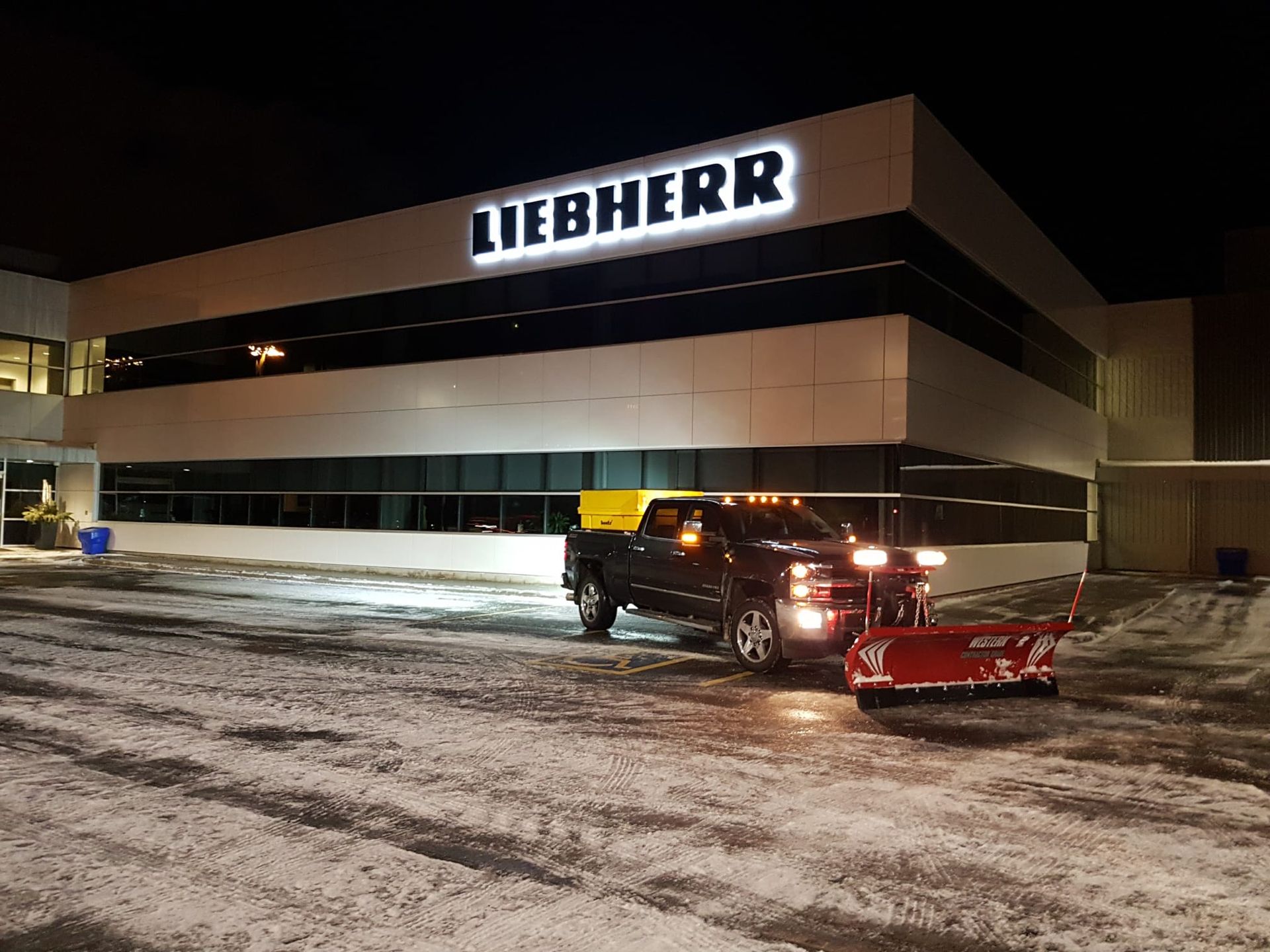 A black truck with a red plow attached is parked in front of a modern building lit up at night.