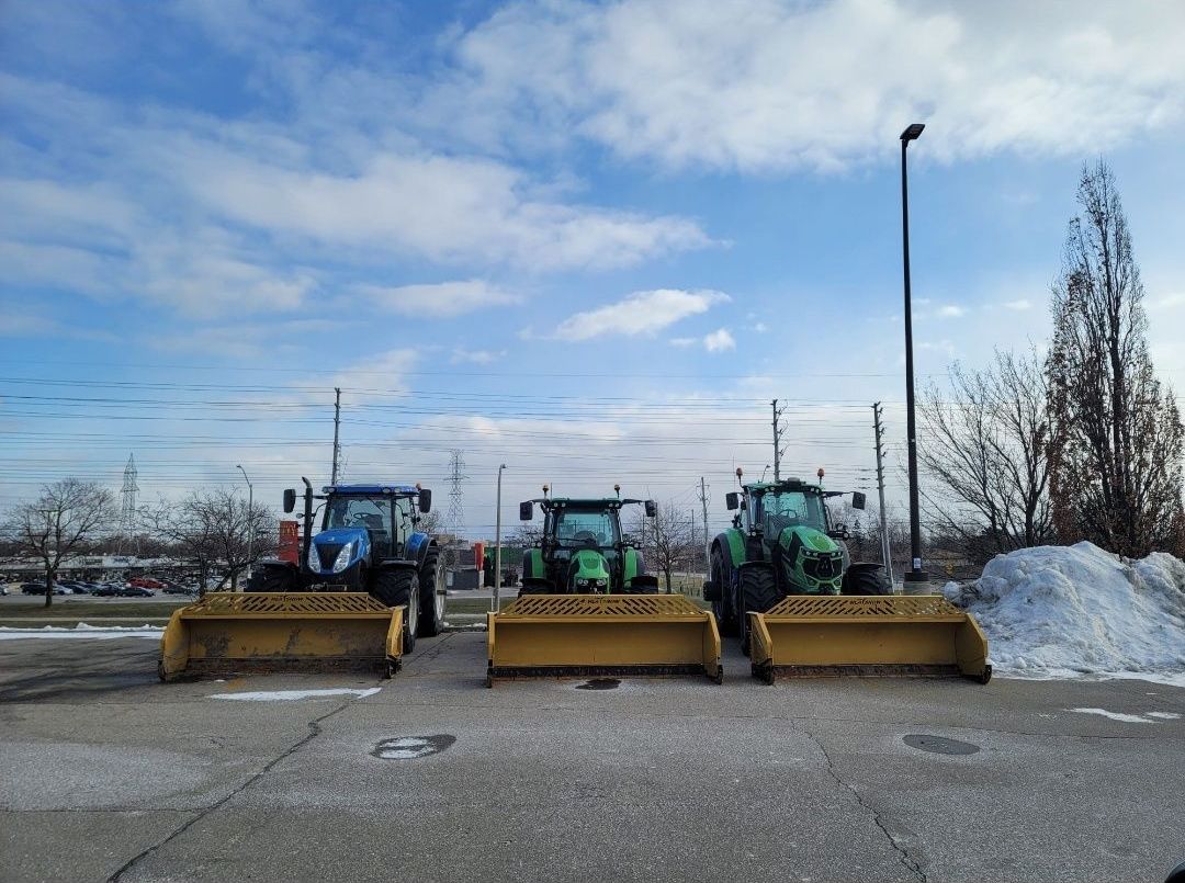 Three tractors equipped with yellow blades parked side by side in a parking lot, with residual snow visible.