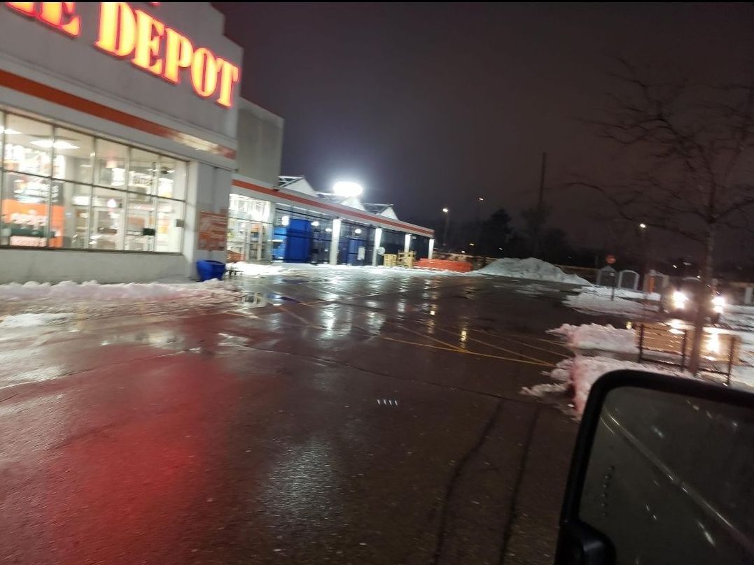 A nighttime parking lot covered in snow and ice in front of an illuminated hardware store, viewed from a vehicle.