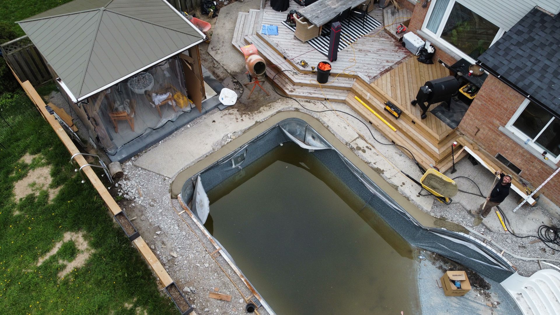 An aerial view shows a backyard with an empty, murky pool, a gazebo, and wooden decking under construction.