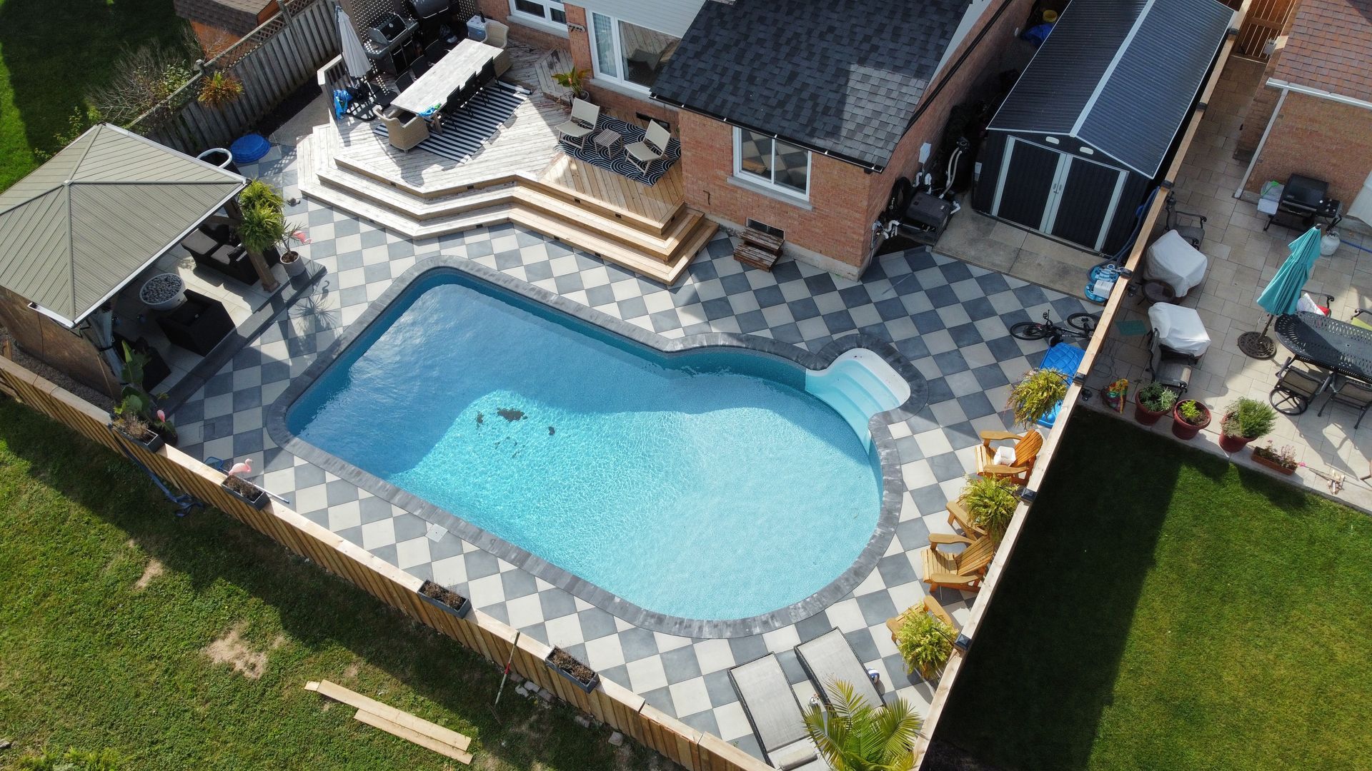 Residential pool with a checkerboard-patterned paved patio and a brick house.