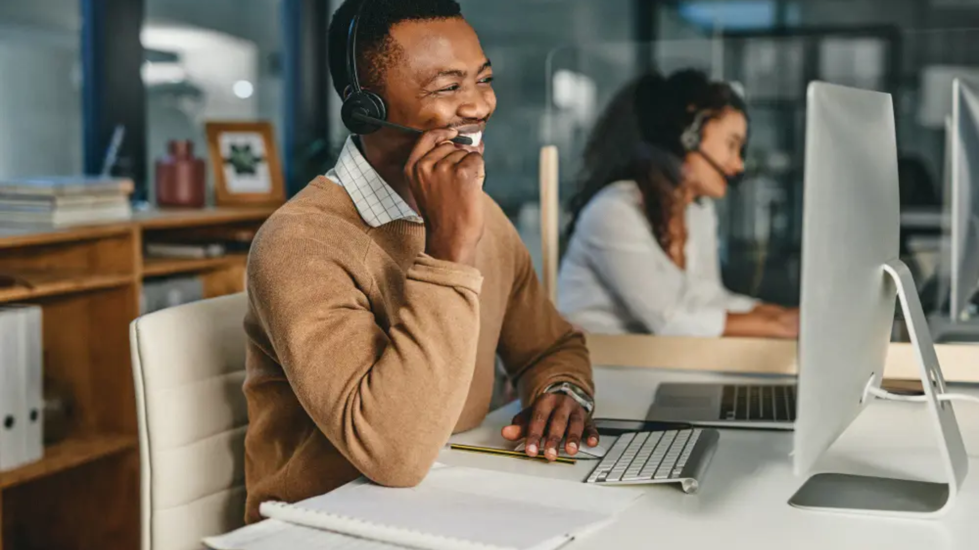 Dos compañeros de trabajo con auriculares puestos en ordenadores de sobremesa en una oficina; uno de ellos sonríe mientras habla por teléfono.