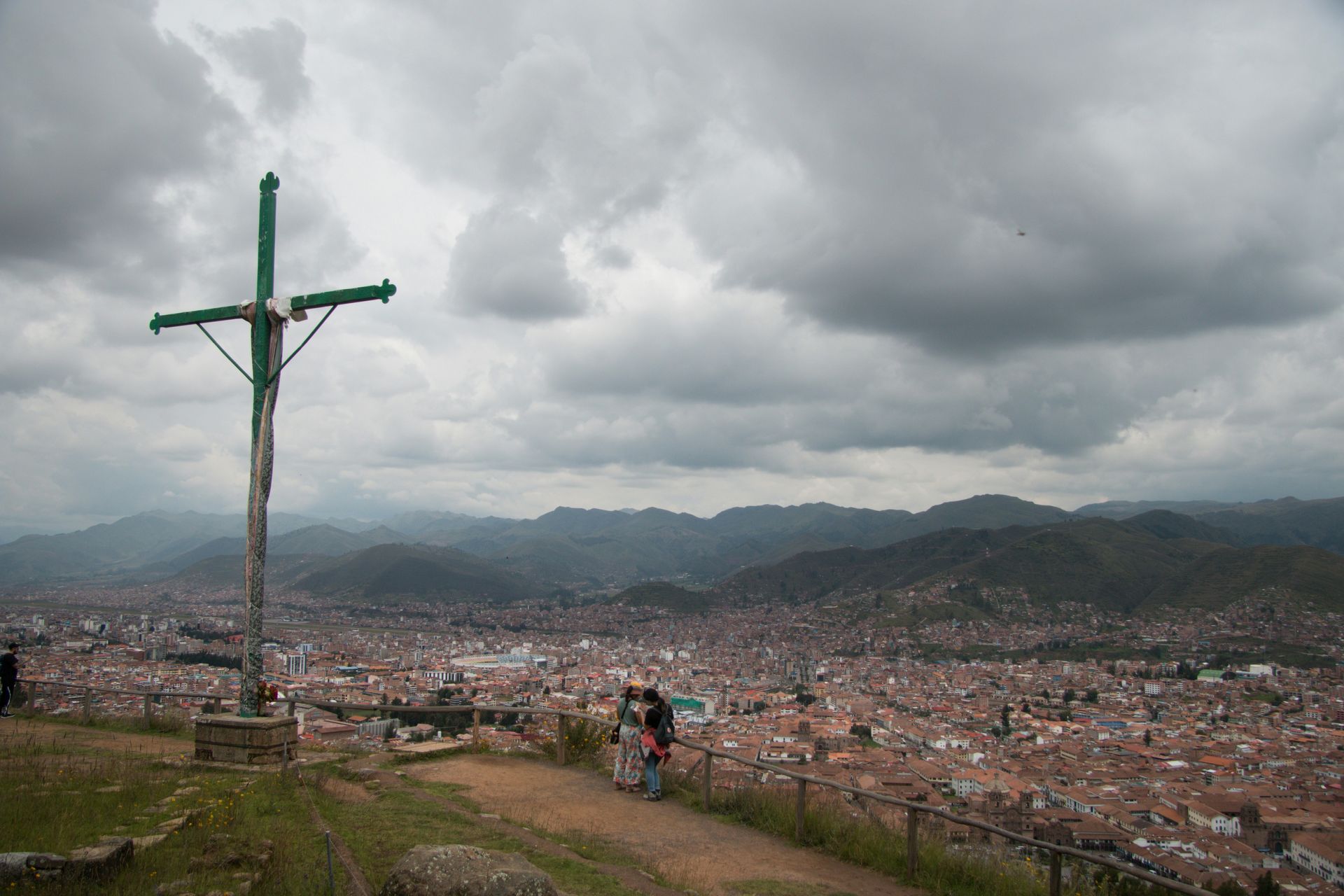 Una gran cruz domina una extensa ciudad bajo cielos oscuros y nublados, con montañas a lo lejos.