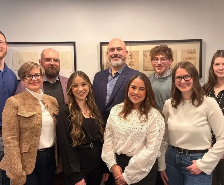 Group of seven colleagues posing indoors in front of framed artwork, smiling at the camera.