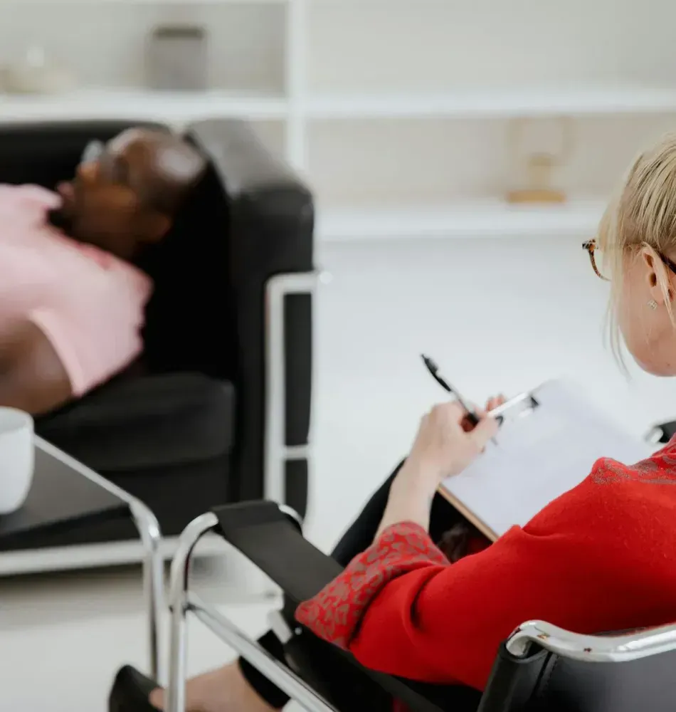 Person in red sweater taking notes while another person reclines in a black chair in a bright office.