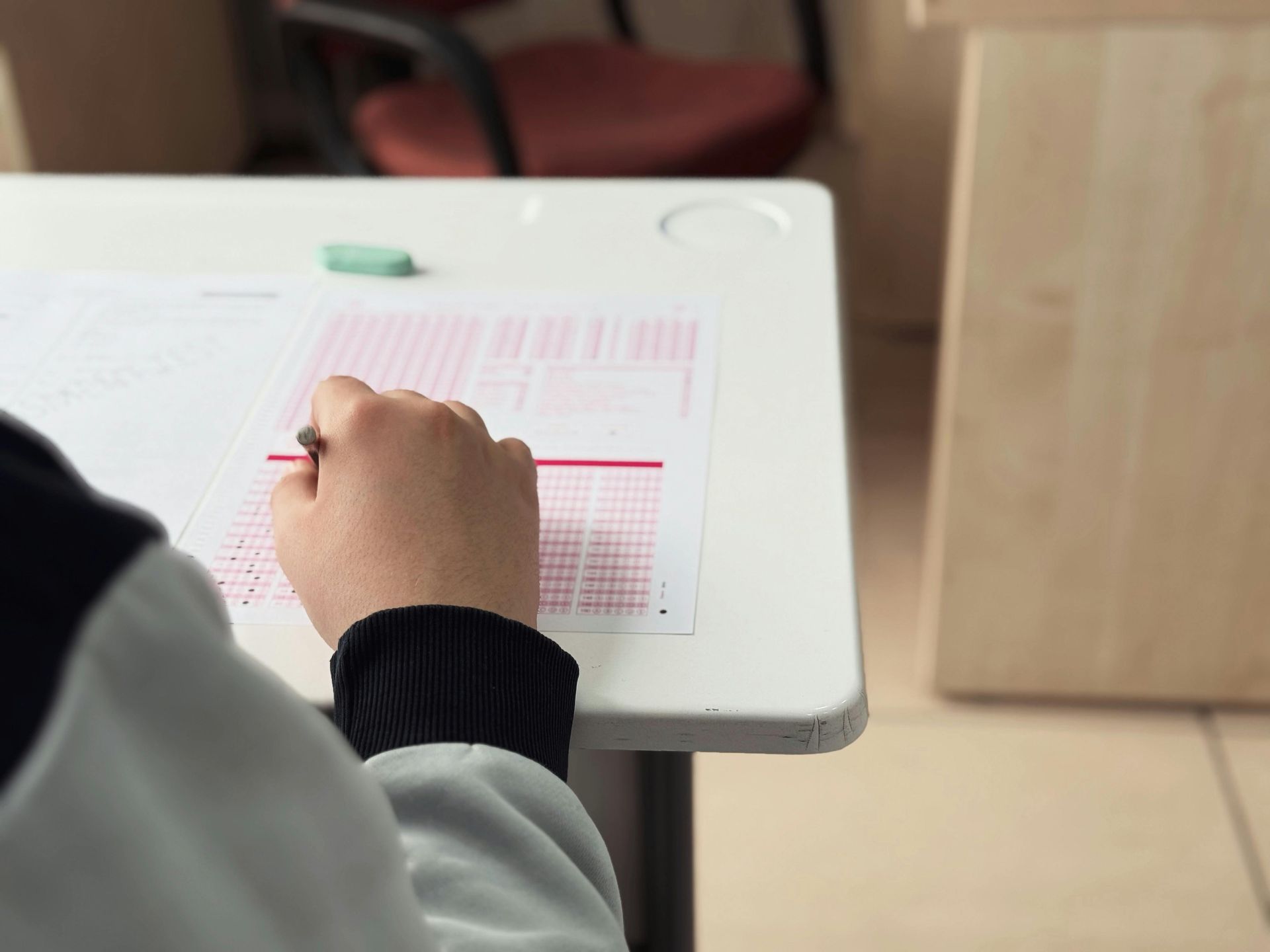 Person writing on a form at a white desk with a red pen, in an office-like room.
