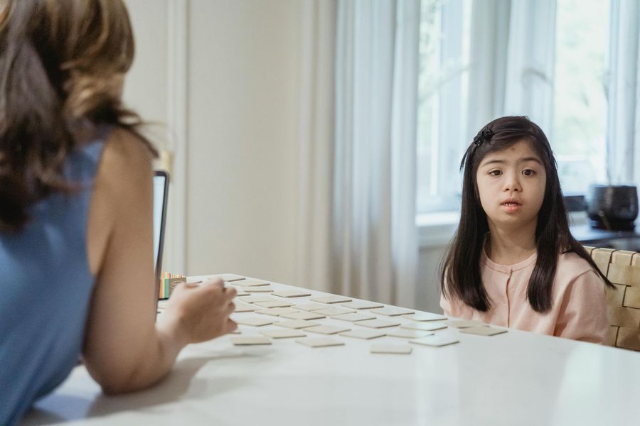 Woman and girl playing a tabletop tile game in a bright room with a window nearby